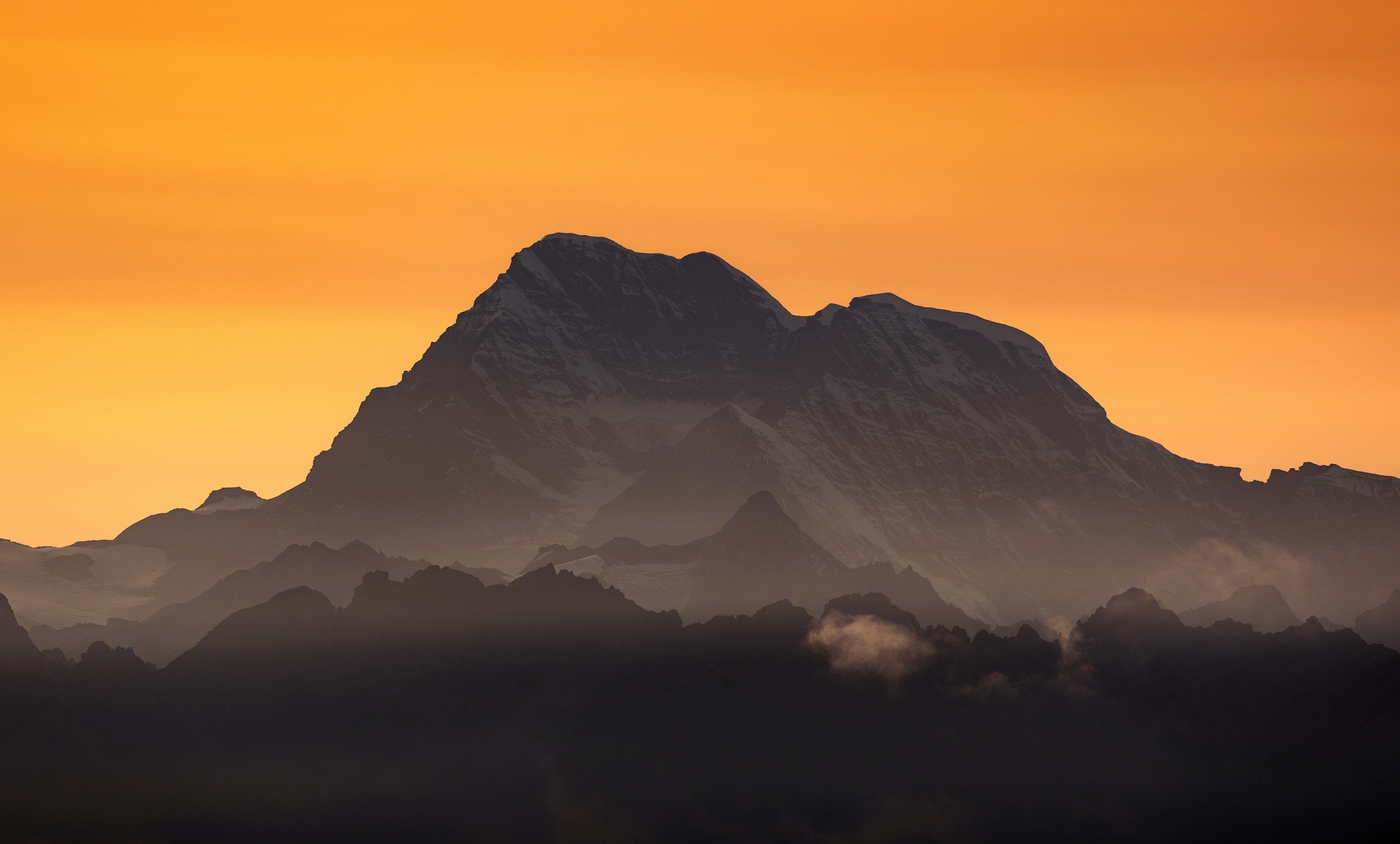 A mountain range at sunset or sunrise with a large prominent peak and misty clouds at the base, under an orange sky.